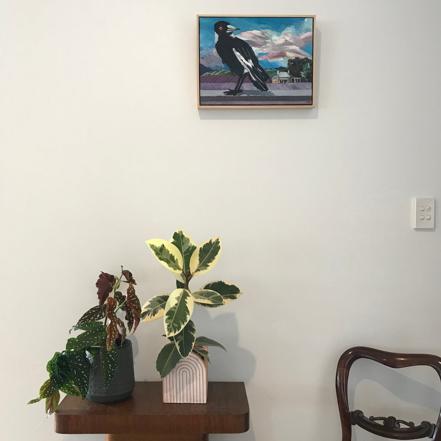 Two potted plants on a wooden table with a framed painting above, against a white wall.