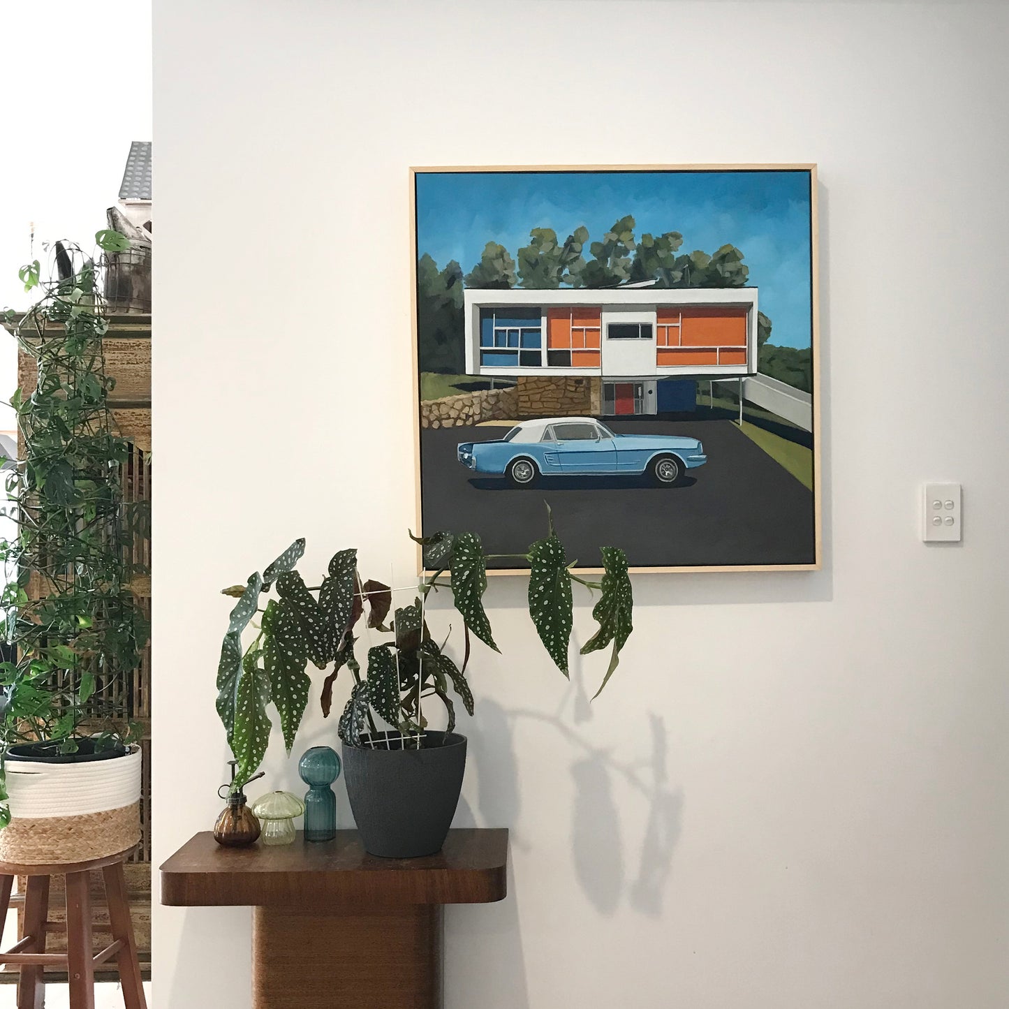 Framed artwork of a modern house with a car parked in front, displayed on a wall above a wooden console table with plants.
