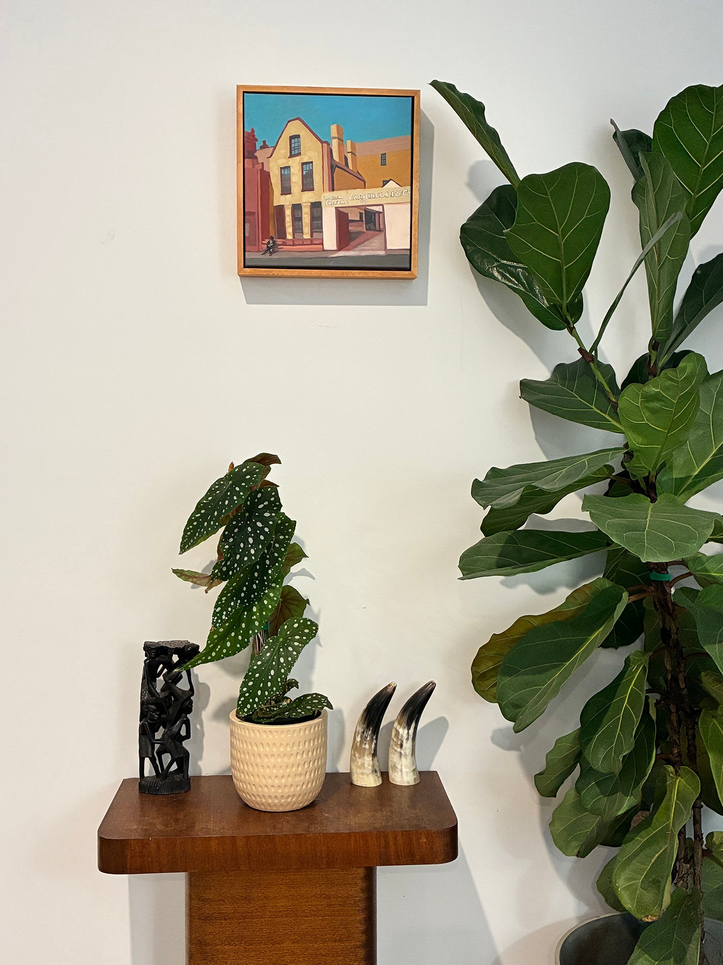 Decorative setup with plants, a small sculpture, and a framed painting on a white wall.