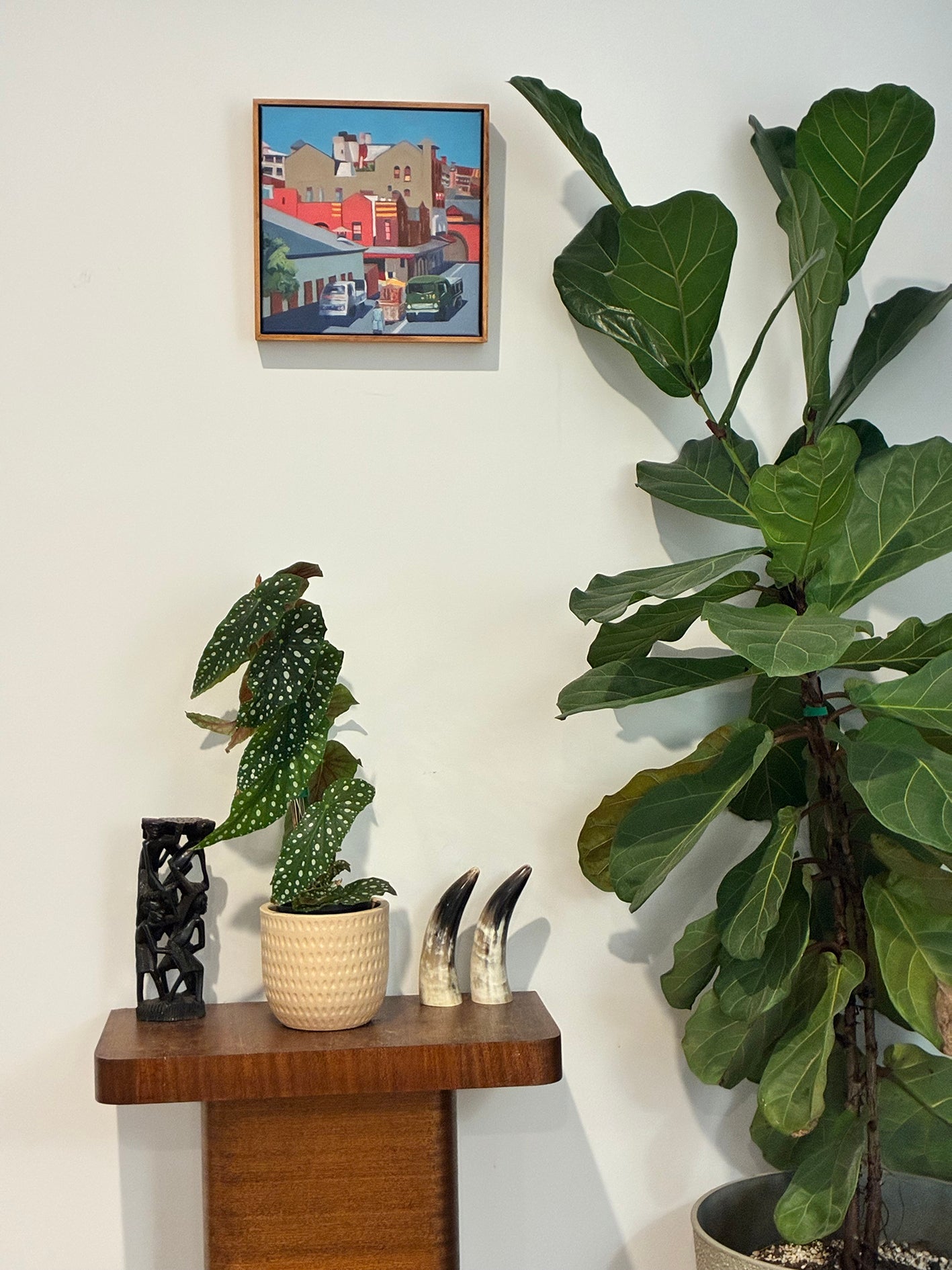 Potted plants on a wooden shelf with a framed picture above against a white wall.