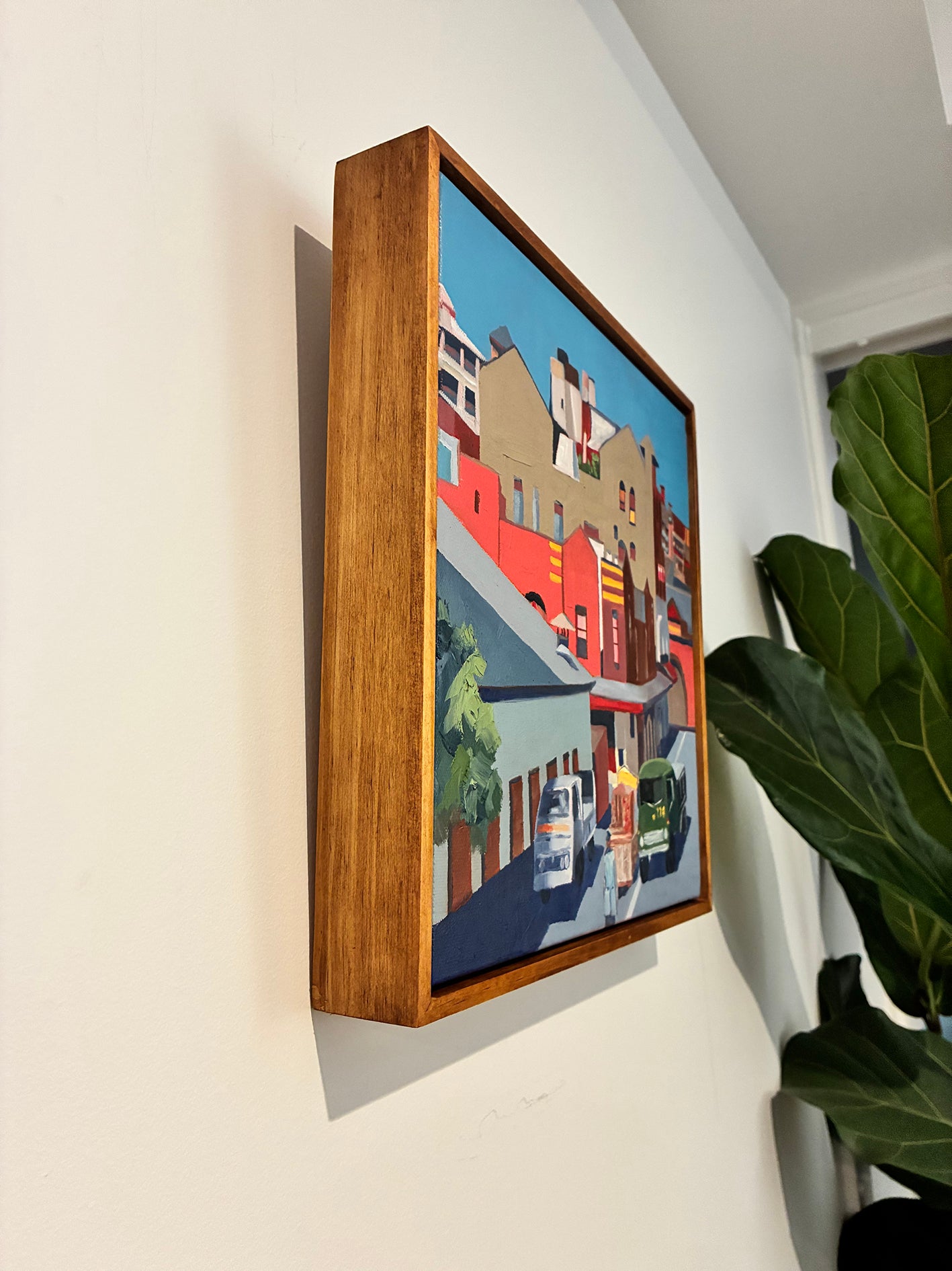 Framed artwork of a cityscape on a wall with a plant to the right.