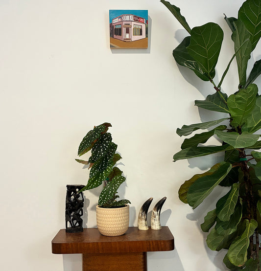 Two potted plants on a wooden shelf with a small painting above them against a white wall.