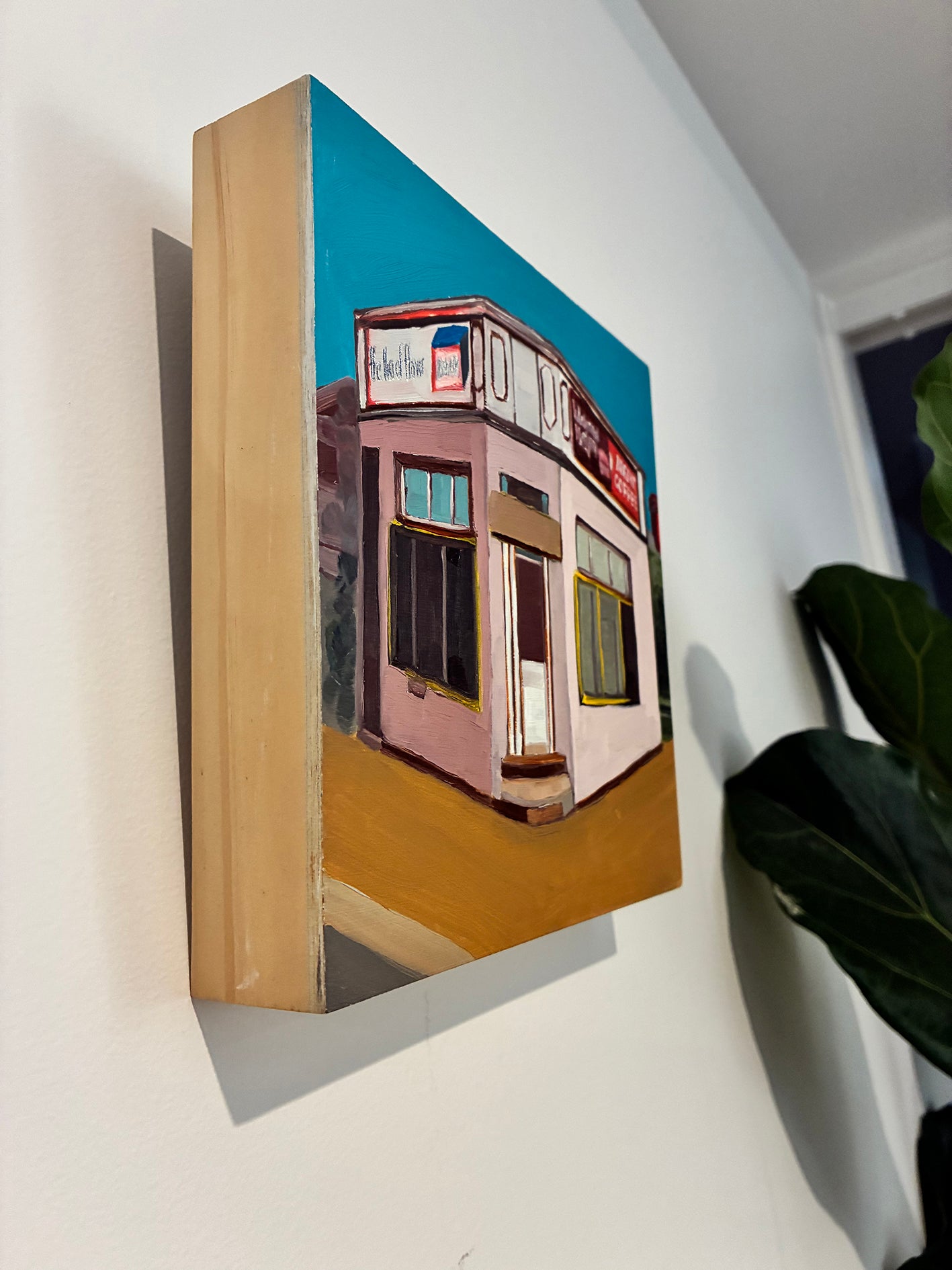Painting of a small store on a wall with a plant in the corner