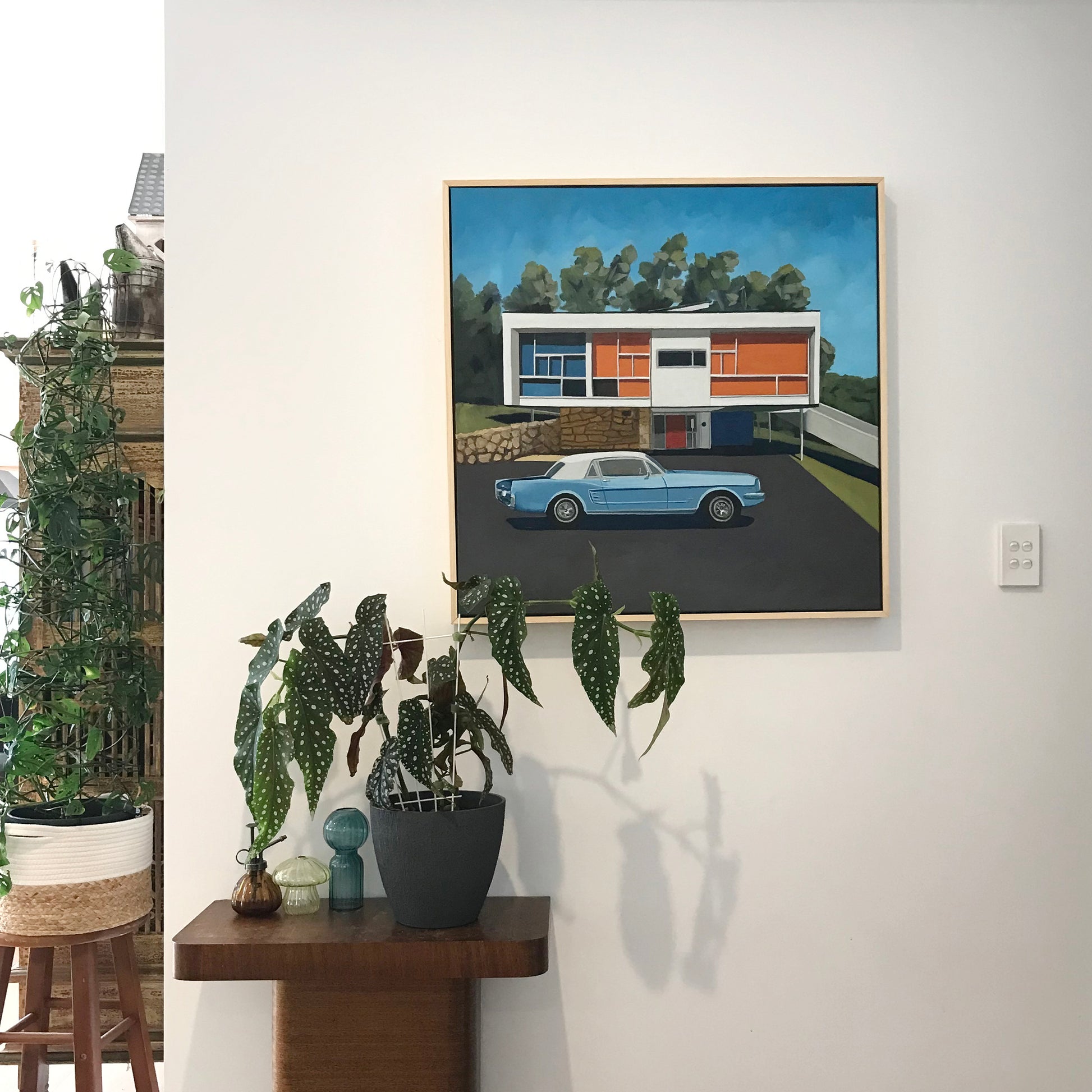 Framed artwork of a modern house with a car parked in front, displayed on a wall above a wooden console table with plants.