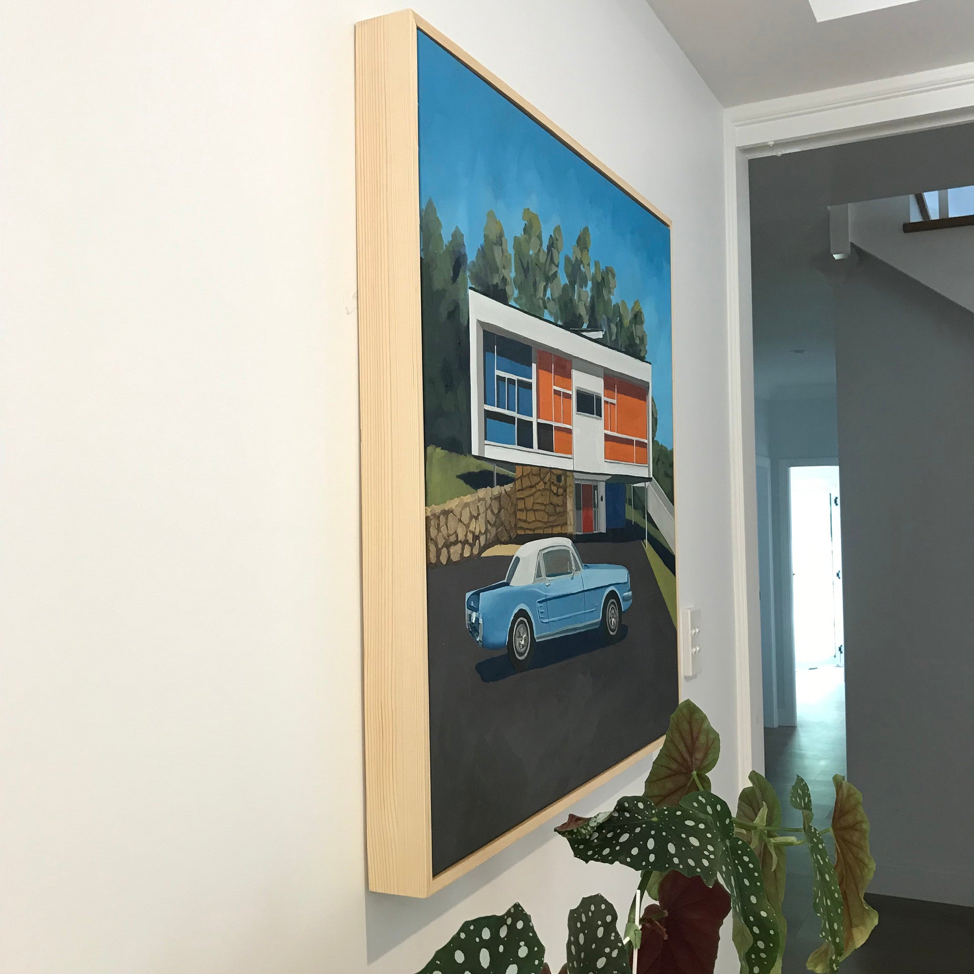 Framed artwork of a modern house with a blue car on a wall in a room.