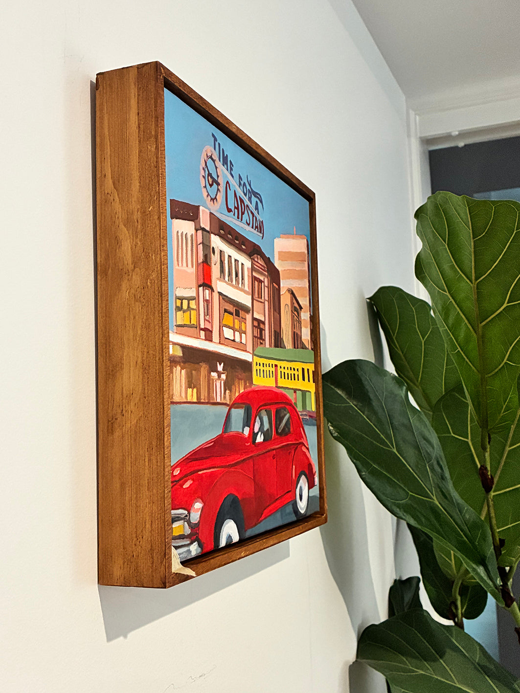 Framed artwork of a red car in front of a building on a wall with a plant in the foreground.