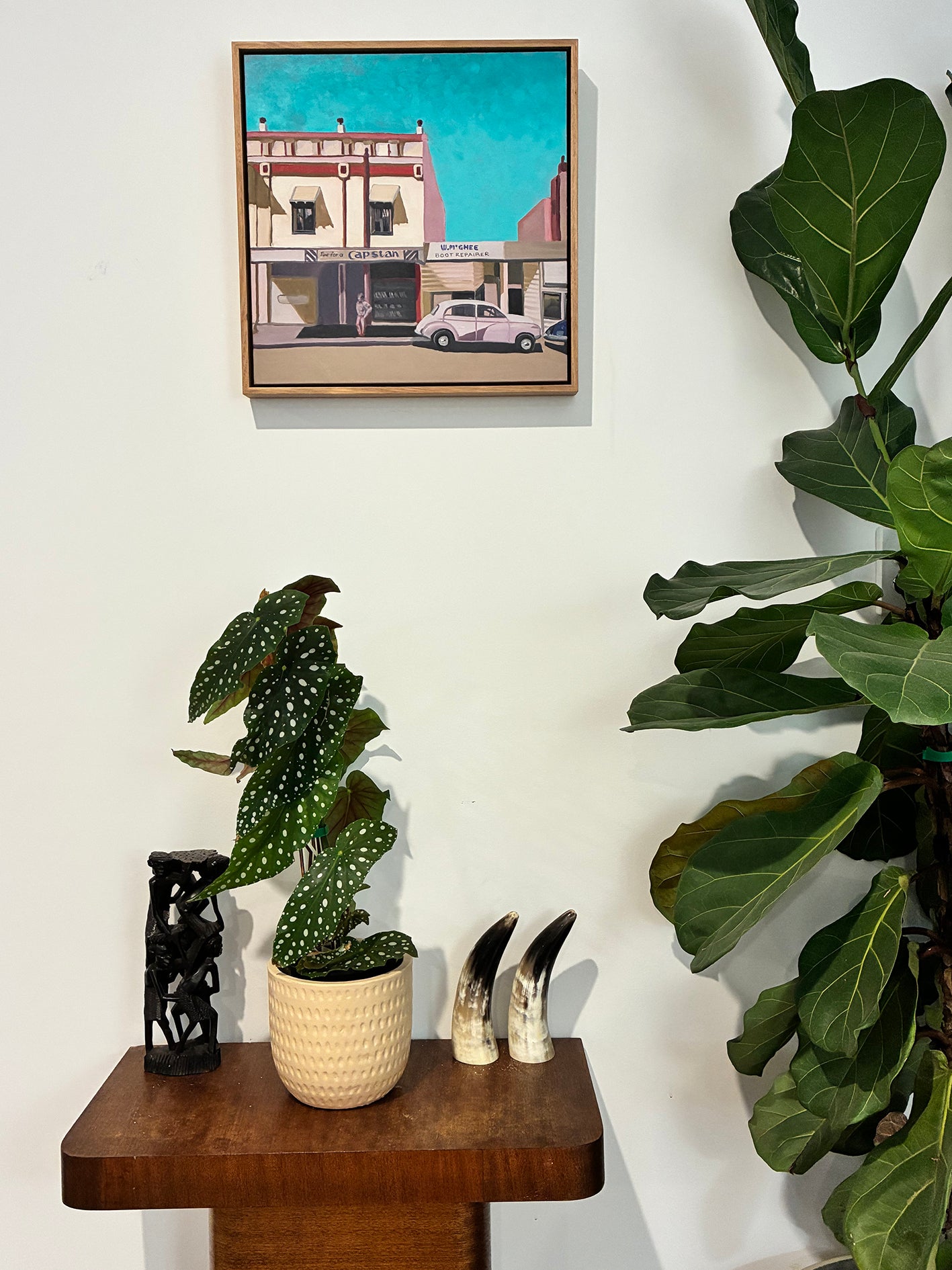 Framed artwork of a city street scene above a small table with decorative items and plants.
