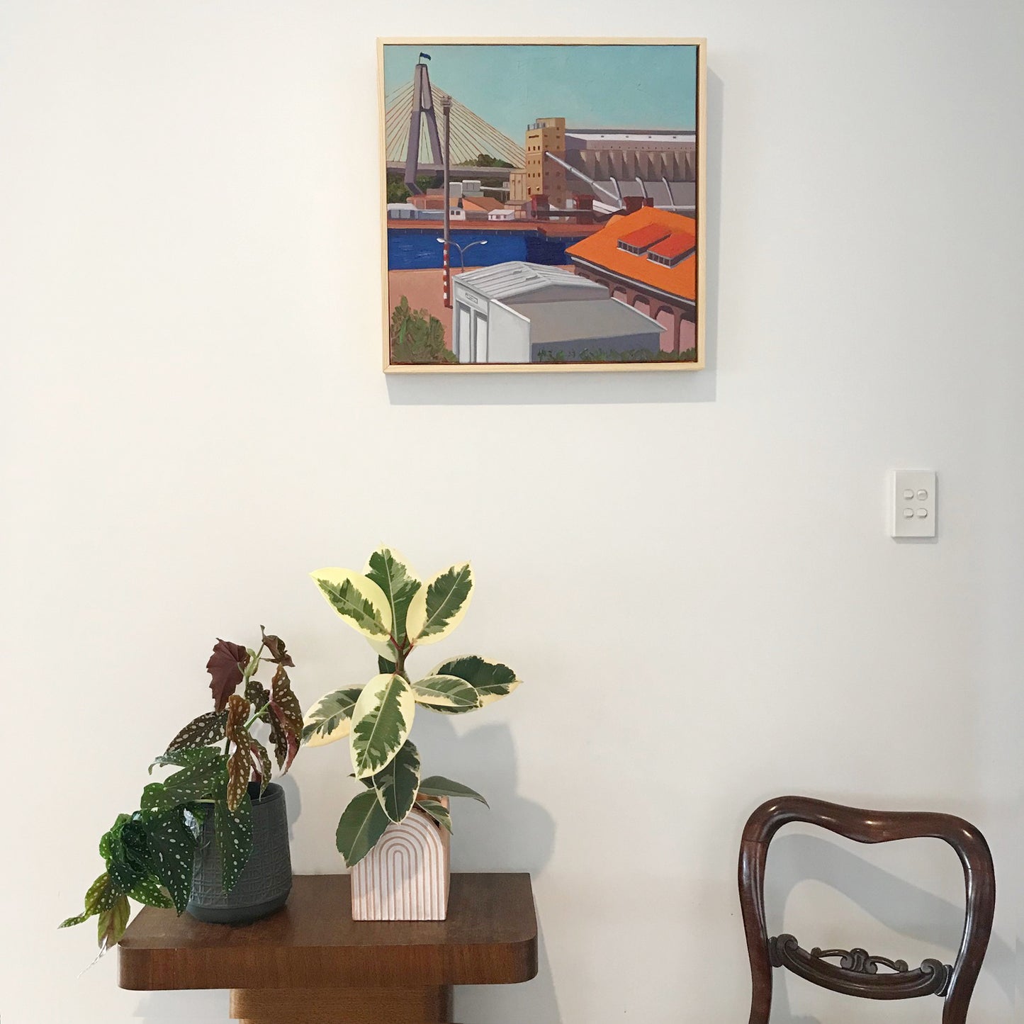 Framed artwork on a wall above a small table with plants and a chair.