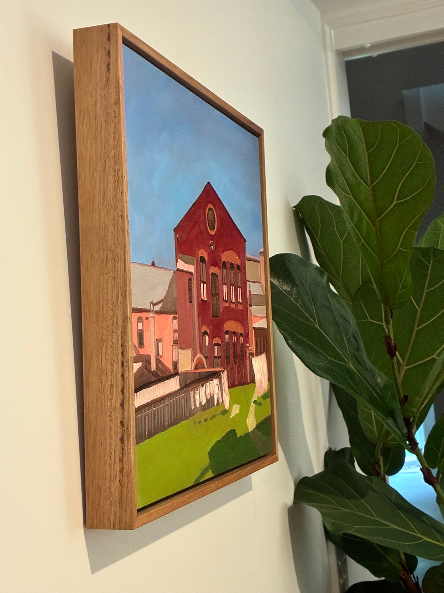 Framed painting of a red brick building on a wall next to a plant